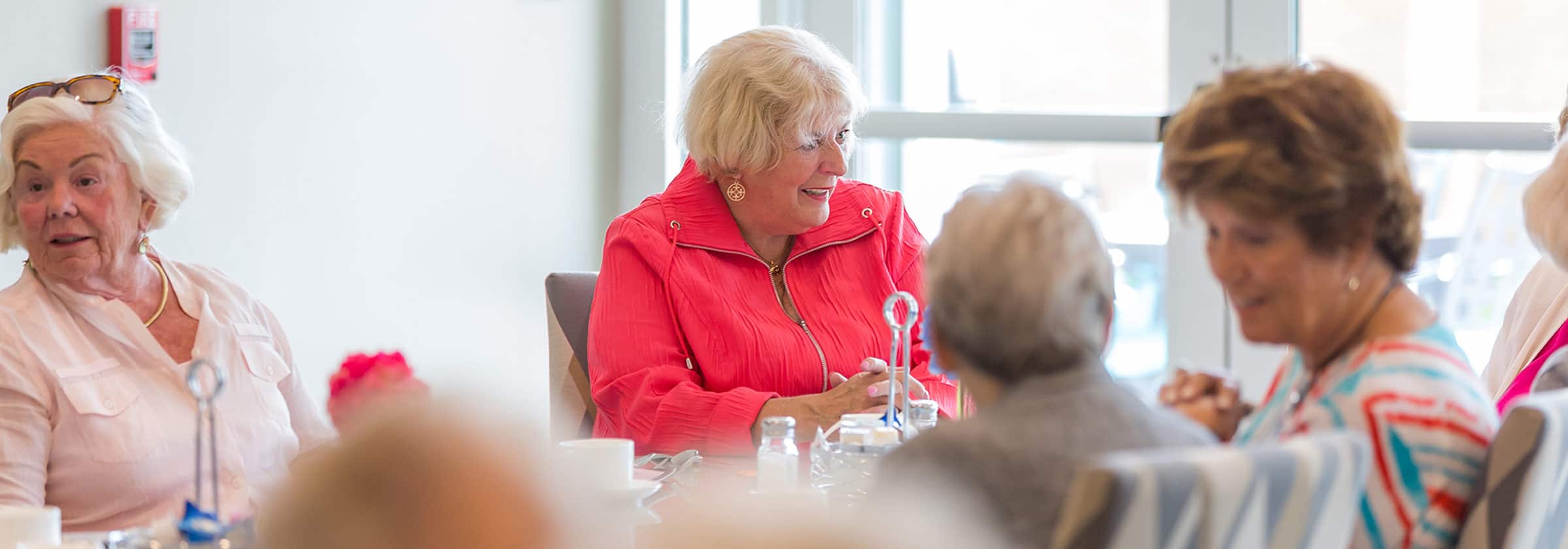 A group of smiling senior women sitting together at a dining table in a senior living facility enjoying a meal