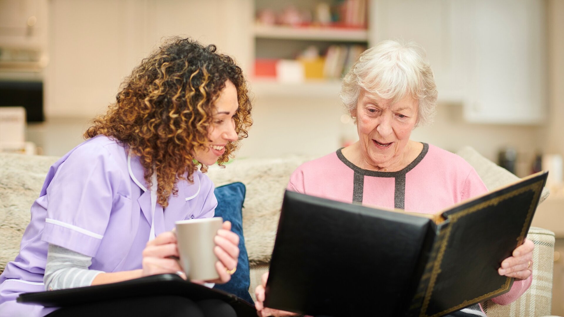 Female memory care resident sitting on a couch in her memory care apartment at Traditions of Medina with a caregiver
