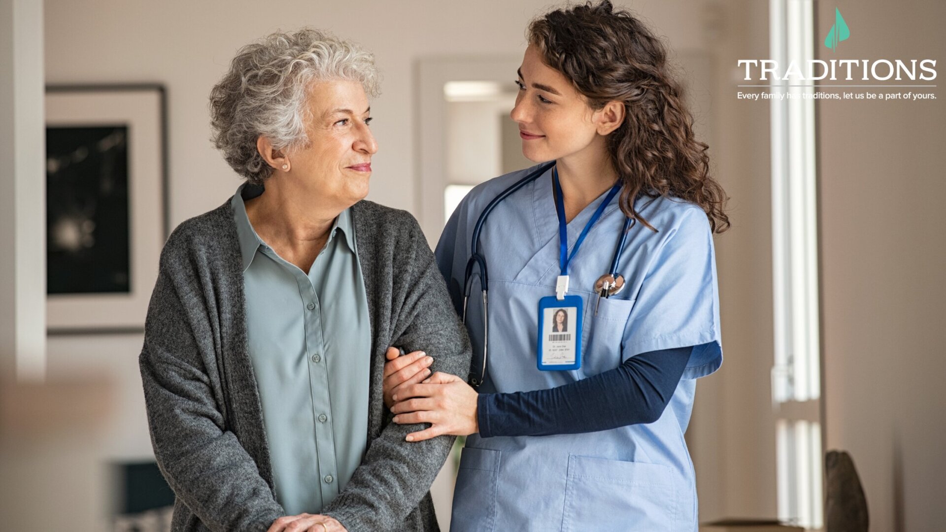 A senior woman with short curly grey hair smiling with her arm linked with her caregivers as they smile at each other with the Traditions Management logo in the top righthand corner of the image
