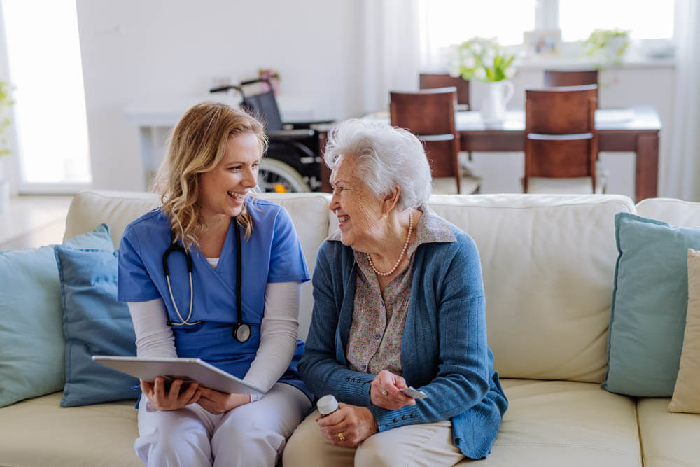 Female nurse sitting on a sofa with a memory care patient, laughing and conversing, at Traditions of Medina in Medina, OH.